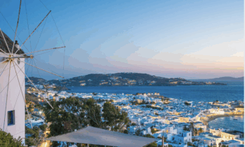 Eine Küstenstadt bei Sonnenuntergang mit weißen Gebäuden, einer Windmühle auf der linken Seite und dem blauen Meer, das sich bis zum Horizont erstreckt. Hügel umrahmen die Stadt, die in weiches, warmes Abendlicht getaucht ist. Das Wort "Geist" erscheint unten rechts.