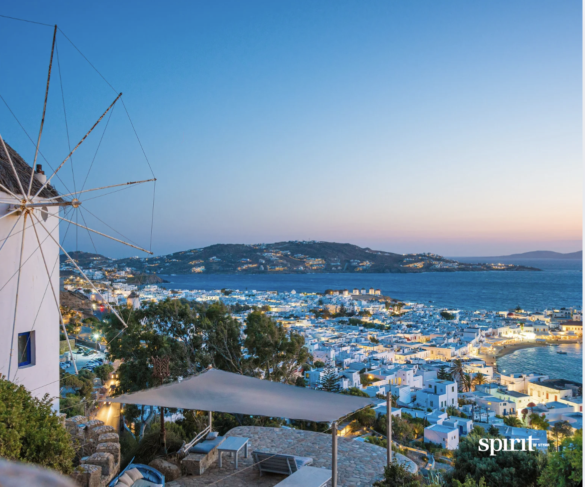 Eine Küstenstadt bei Sonnenuntergang mit weißen Gebäuden, einer Windmühle auf der linken Seite und dem blauen Meer, das sich bis zum Horizont erstreckt. Hügel umrahmen die Stadt, die in weiches, warmes Abendlicht getaucht ist. Das Wort "Geist" erscheint unten rechts.