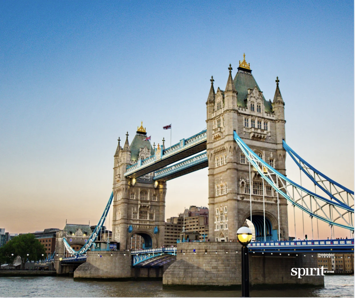 Die Tower Bridge in London mit ihren zwei Türmen und blauen Hängebrücken überspannt die Themse bei Sonnenuntergang. Der Himmel ist klar, und das Wort "Spirit" erscheint in der unteren rechten Ecke.
