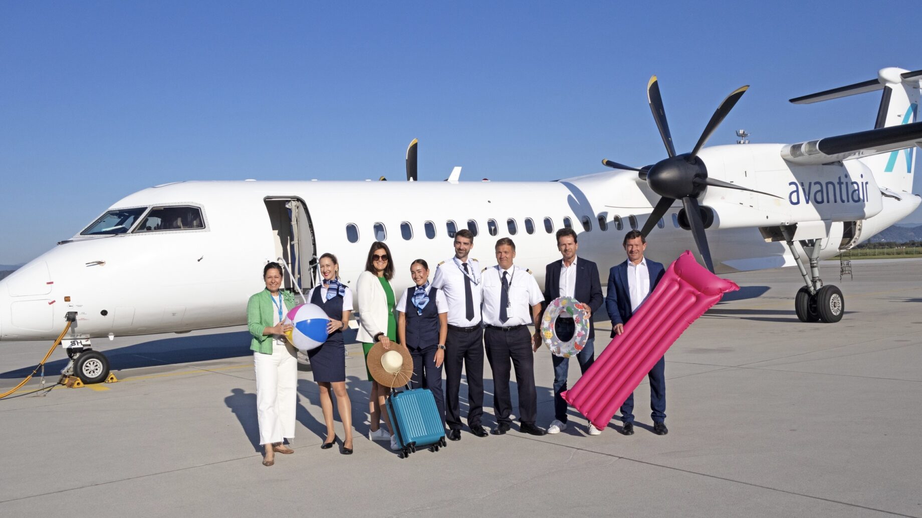 Acht Personen, einige in Uniform, andere in Geschäftskleidung, stehen lächelnd vor einem Avantiair-Flugzeug auf dem Rollfeld eines sonnigen Flughafens und halten Urlaubsgegenstände wie einen Strandball, einen Hut, einen Koffer, ein Floß und eine Luftmatratze.