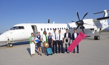 Acht Personen, einige in Uniform, andere in Geschäftskleidung, stehen lächelnd vor einem Avantiair-Flugzeug auf dem Rollfeld eines sonnigen Flughafens und halten Urlaubsgegenstände wie einen Strandball, einen Hut, einen Koffer, ein Floß und eine Luftmatratze.