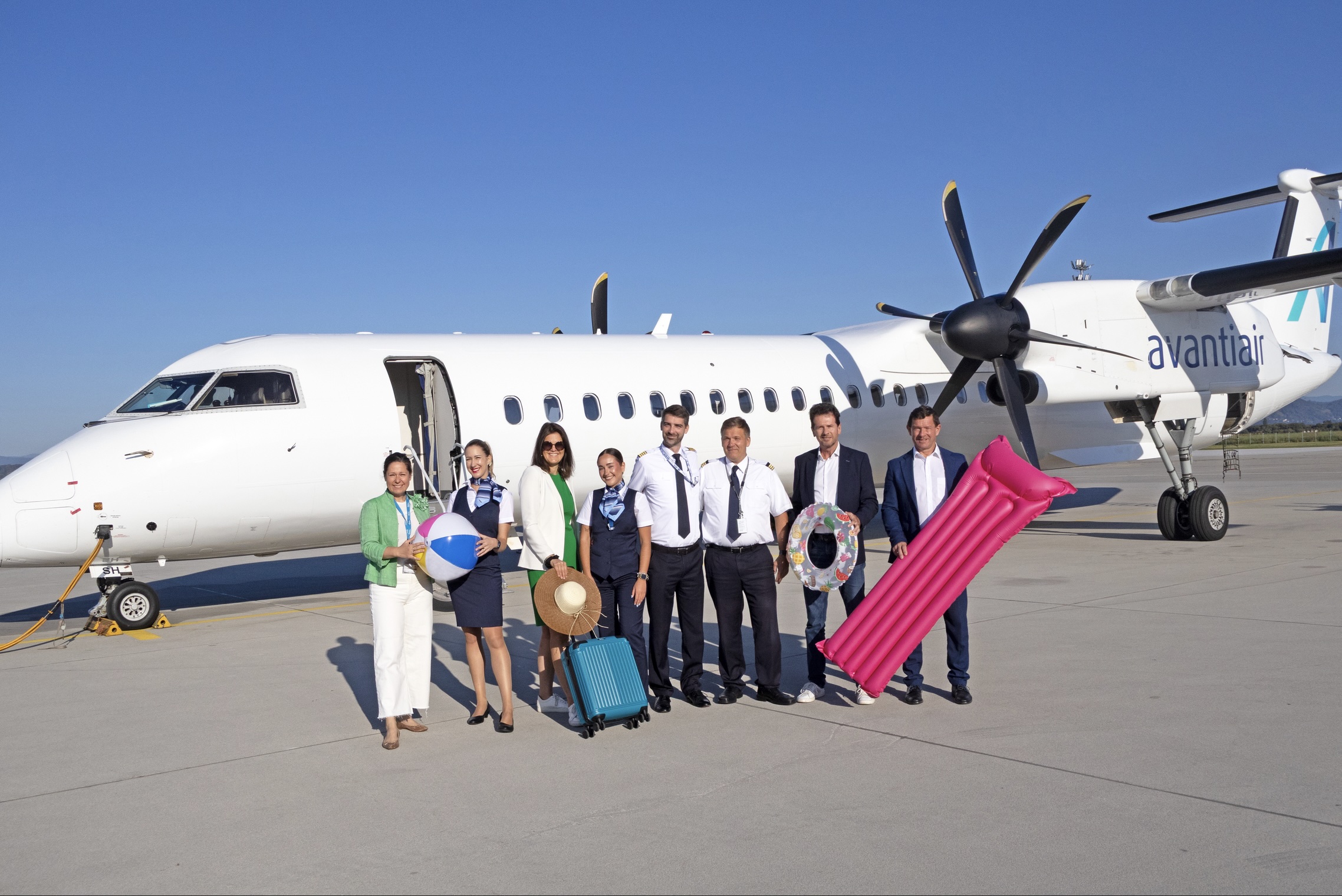 Acht Personen, einige in Uniform, andere in Geschäftskleidung, stehen lächelnd vor einem Avantiair-Flugzeug auf dem Rollfeld eines sonnigen Flughafens und halten Urlaubsgegenstände wie einen Strandball, einen Hut, einen Koffer, ein Floß und eine Luftmatratze.