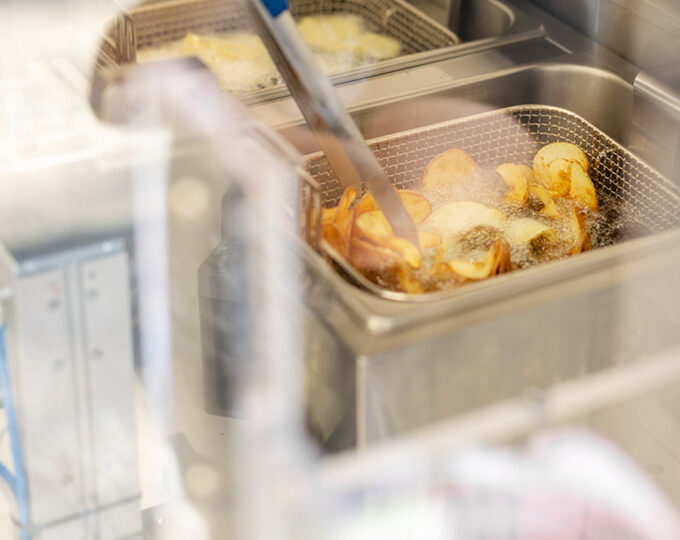 Eine Nahaufnahme von Kartoffelscheiben, die in einem Frittierkorb frittiert werden, wobei eine Zange das Essen umrührt. Die Bedienknöpfe der Metallfritteuse sind rechts zu sehen. Durch eine Glas- oder Kunststoffbarriere aufgenommen, deutet diese Szene auf nachhaltiges Bauen in Großküchen hin.