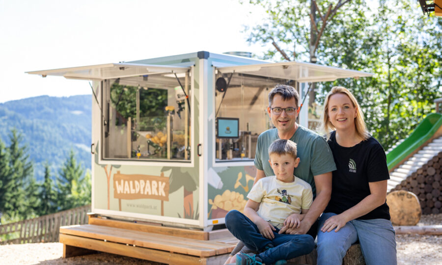 Ein Mann, eine Frau und ein kleiner Junge sitzen lächelnd vor einem kleinen Kiosk mit der Aufschrift "Waldpark", der an einem sonnigen Tag inmitten von Bäumen und Hügeln Immobilien und Bauen präsentiert und eine Atmosphäre der Nachhaltigkeit widerspiegelt.