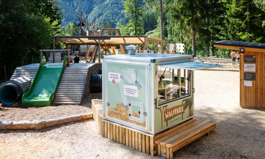 Ein kleiner grüner Imbisskiosk, der mit dem Schwerpunkt Nachhaltigkeit gebaut wurde, steht vor einem Waldspielplatz mit Rutschen und Klettergerüsten, umgeben von Bäumen und Bergen im Hintergrund.