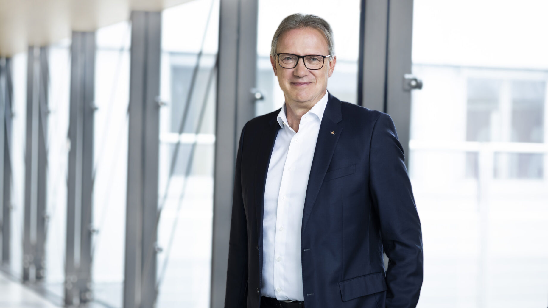 Ein Mann mit Brille und dunklem Anzug steht in einem hellen, modernen Flur mit großen Fenstern und lächelt in die Kamera - das spiegelt den Geist der Nachhaltigkeit in modernen Immobilien wider.