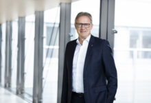 Ein Mann mit Brille und dunklem Anzug steht in einem hellen, modernen Flur mit großen Fenstern und lächelt in die Kamera - das spiegelt den Geist der Nachhaltigkeit in modernen Immobilien wider.