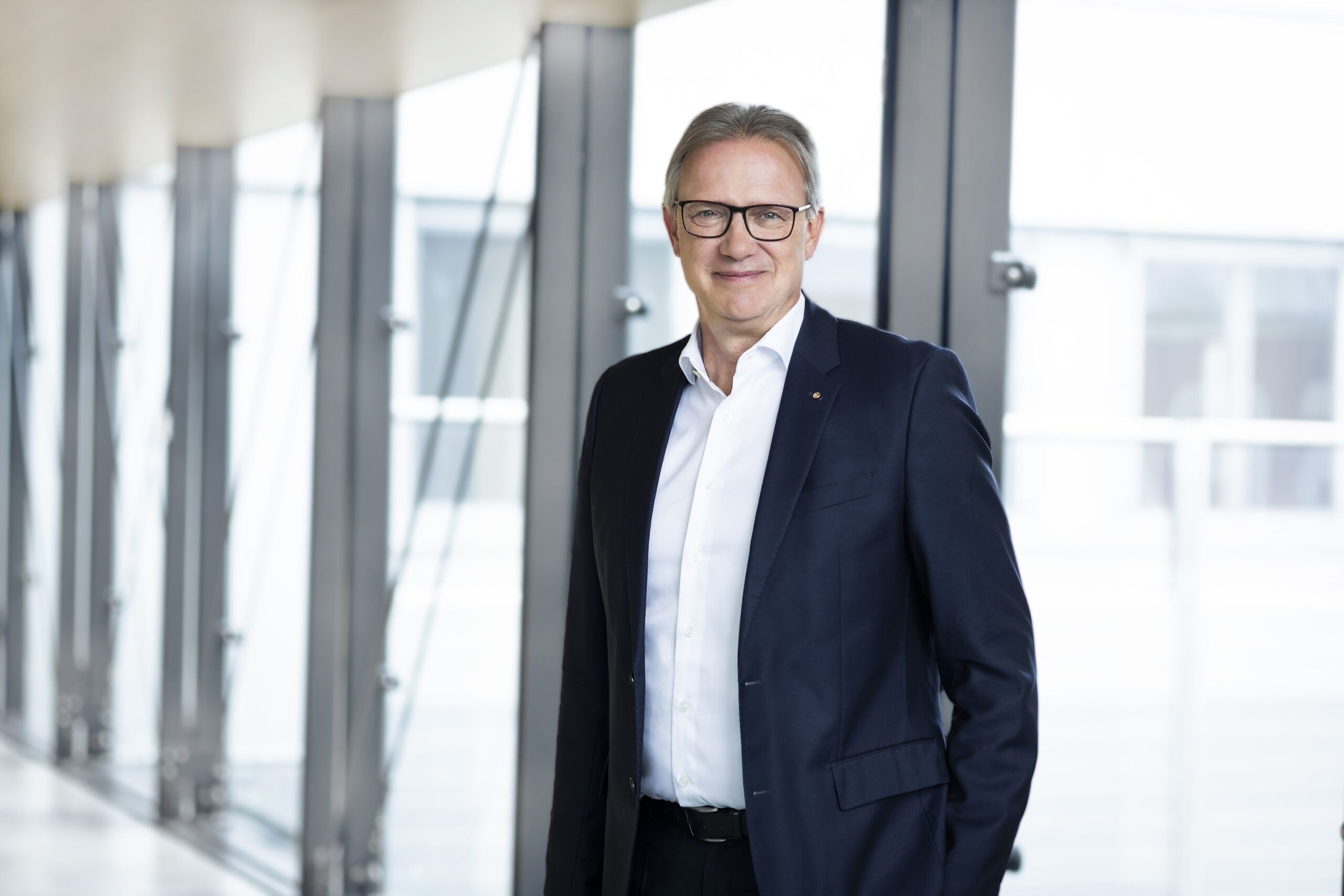 Ein Mann mit Brille und dunklem Anzug steht in einem hellen, modernen Flur mit großen Fenstern und lächelt in die Kamera - das spiegelt den Geist der Nachhaltigkeit in modernen Immobilien wider.