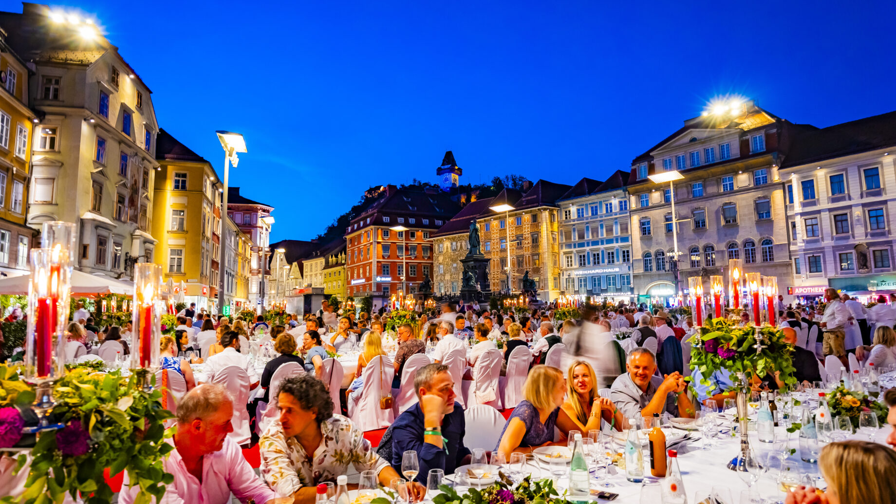 Ein lebhaftes Abendessen unter freiem Himmel findet in der Abenddämmerung auf einem europäischen Stadtplatz statt, wo viele Menschen an langen Tischen unter dekorativen Lichtern sitzen, umgeben von farbenfrohen historischen Gebäuden.