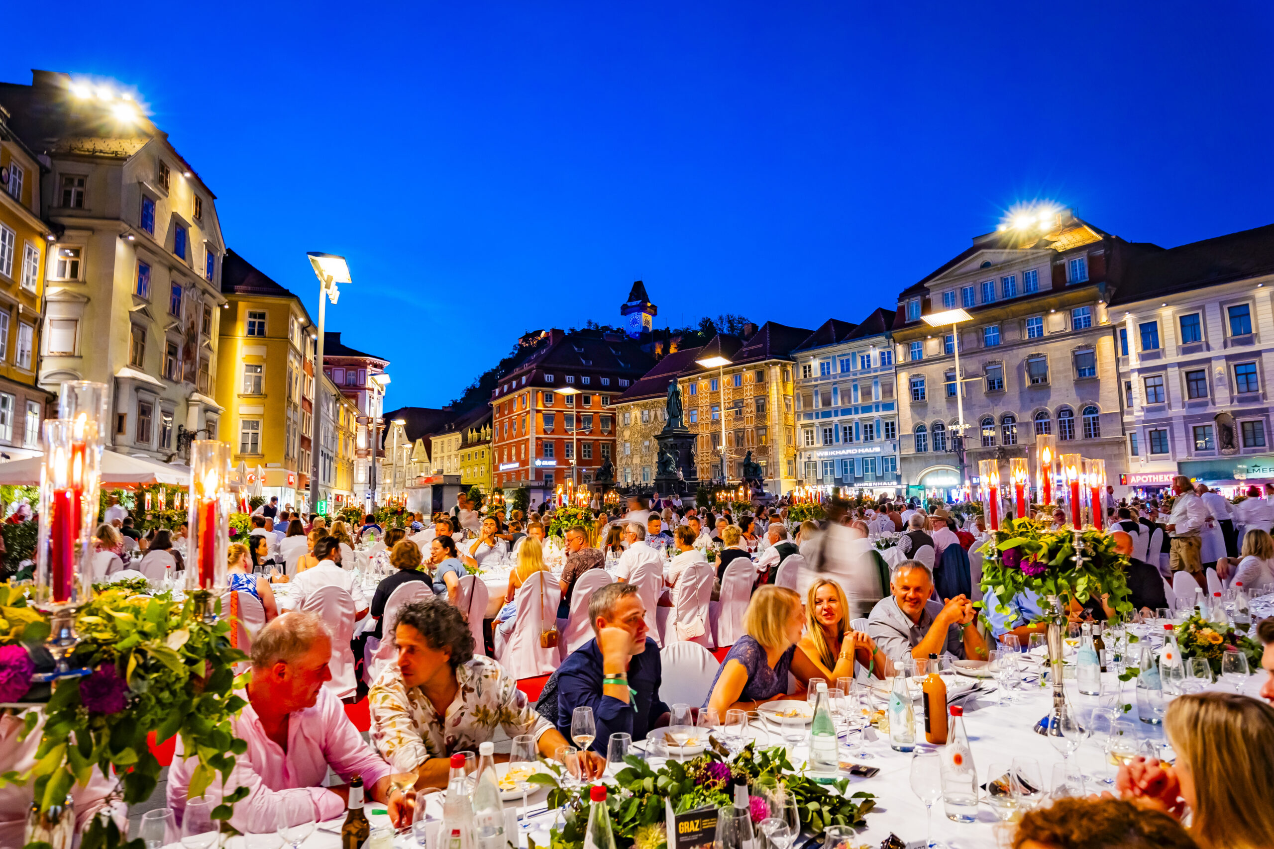Ein lebhaftes Abendessen unter freiem Himmel findet in der Abenddämmerung auf einem europäischen Stadtplatz statt, wo viele Menschen an langen Tischen unter dekorativen Lichtern sitzen, umgeben von farbenfrohen historischen Gebäuden.