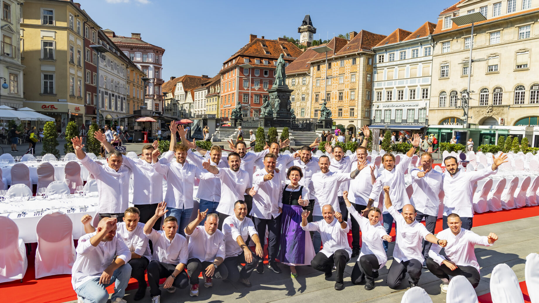 Eine Gruppe von Köchen in weißen Jacken posiert und feiert gemeinsam auf einem europäischen Stadtplatz, umgeben von langen Tischen mit weißen Tischtüchern und historischen, farbenfrohen Gebäuden unter einem sonnigen blauen Himmel.