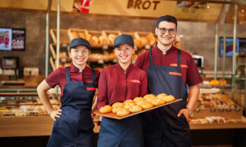 Drei lächelnde Bäckereiangestellte in Uniform stehen vor den Regalen voller Brot. Die Bäckerei "Brot" setzt auf regionale Qualität und ist stolz darauf, den Steiermark Markt mit jeder frischen Semmel als schmackhafte Investition in den lokalen Geschmack zu unterstützen.