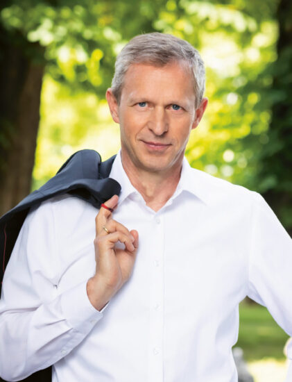 Ein Mann mittleren Alters mit grauem Haar steht im Freien, trägt ein weißes Hemd und eine dunkle Hose, hält mit einer Hand eine Jacke über die Schulter und stützt die andere Hand auf die Hüfte. Im Hintergrund sind Bäume und Grünflächen zu sehen.