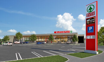 Ein moderner EUROSPAR-Supermarkt mit einer großen Glasfassade präsentiert nachhaltiges Bauen, einen größtenteils leeren Parkplatz, mehrere geparkte Autos, Bäume und ein hohes Schild mit dem Namen des Geschäfts und Parkinformationen unter einem blauen Himmel mit einigen Wolken.