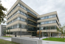 Ein modernes, mehrstöckiges Bürogebäude mit horizontalen beigen und schwarzen Linien, großen Fenstern und Bänken im Freien steht inmitten von grünem Gras und Bäumen unter einem wolkenverhangenen Himmel - das Gebäude Peter-Tunner-Gasse Tragwerke geschafft.