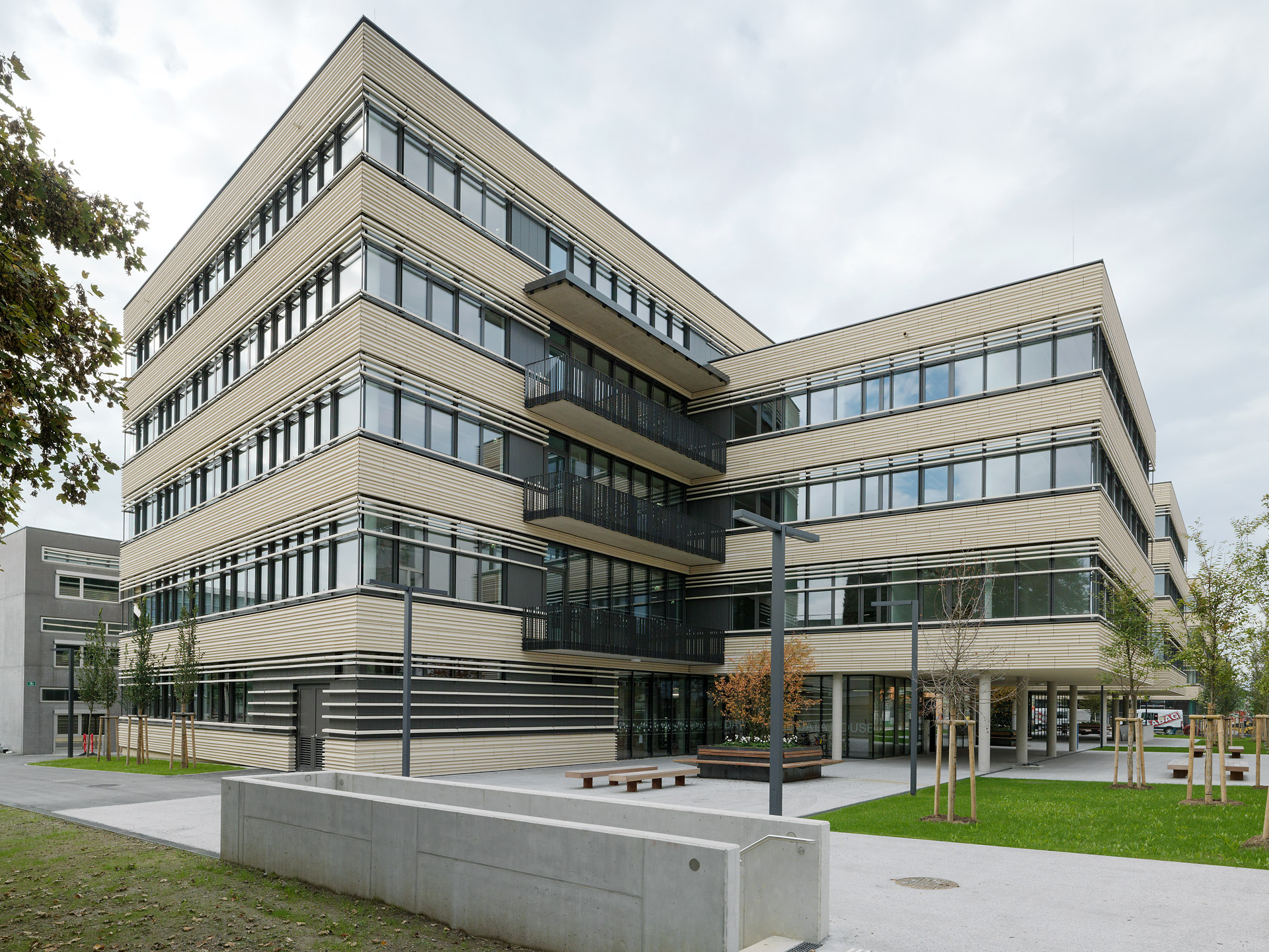 Ein modernes, mehrstöckiges Bürogebäude mit horizontalen beigen und schwarzen Linien, großen Fenstern und Bänken im Freien steht inmitten von grünem Gras und Bäumen unter einem wolkenverhangenen Himmel - das Gebäude Peter-Tunner-Gasse Tragwerke geschafft.
