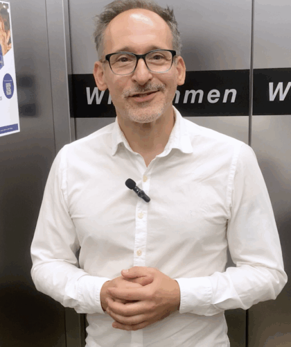 Ein Mann mit Brille und kurzem Bart, der ein weißes Hemd mit einem Ansteckmikrofon trägt, steht vor einer Metallwand mit einem Schild, auf dem teilweise "Willkommen" steht. Er lächelt und fasst sich in die Hände.