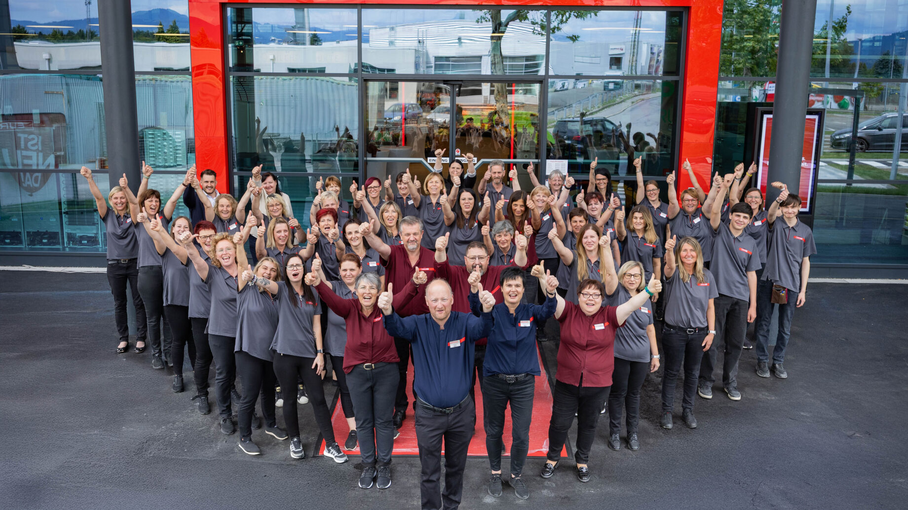Eine große Gruppe lächelnder Menschen in passenden Hemden steht vor einem Glasgebäude und posiert mit erhobenen Armen innerhalb eines roten rechteckigen Rahmens. Die Szene spiegelt Teamgeist im Bauen und Immobilien wider, mit Bäumen und geparkten Autos im Hintergrund.
