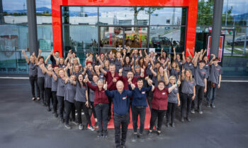 Eine große Gruppe lächelnder Menschen in passenden Hemden steht vor einem Glasgebäude und posiert mit erhobenen Armen innerhalb eines roten rechteckigen Rahmens. Die Szene spiegelt Teamgeist im Bauen und Immobilien wider, mit Bäumen und geparkten Autos im Hintergrund.