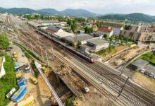 Luftaufnahme einer Eisenbahnbaustelle in der Steiermark mit mehreren Gleisen, einem rot-weißen Zug, nahegelegenen Gebäuden, Baufahrzeugen und grünen Hügeln, die das regionale Wachstum und die zunehmende Zuwanderung der Bevölkerung widerspiegeln.