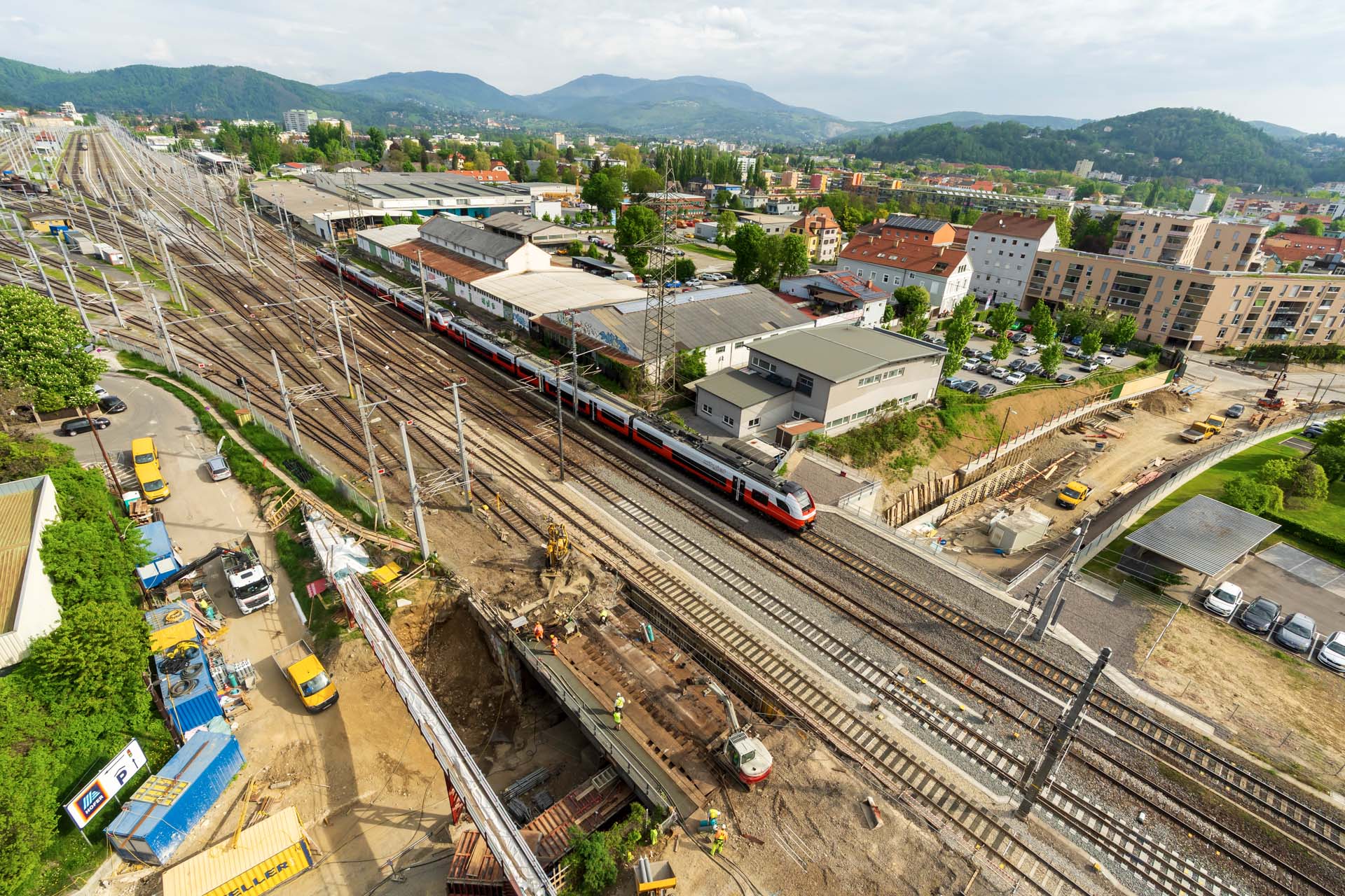 Luftaufnahme einer Eisenbahnbaustelle in der Steiermark mit mehreren Gleisen, einem rot-weißen Zug, nahegelegenen Gebäuden, Baufahrzeugen und grünen Hügeln, die das regionale Wachstum und die zunehmende Zuwanderung der Bevölkerung widerspiegeln.
