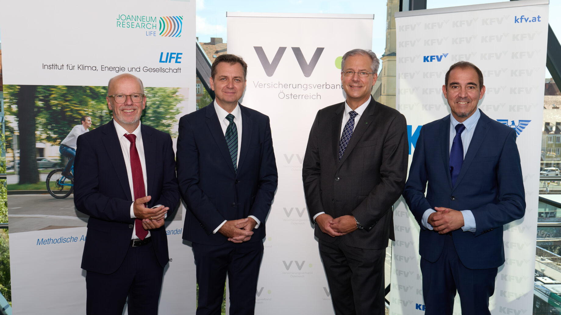 Vier Männer in Business-Kleidung stehen lächelnd vor Bannern von Joanneum Research LIFE, Versicherungsverband Österreich, KFV.at und der langen Tafel Graz in einem modernen Glasgebäude mit Stadtblick.