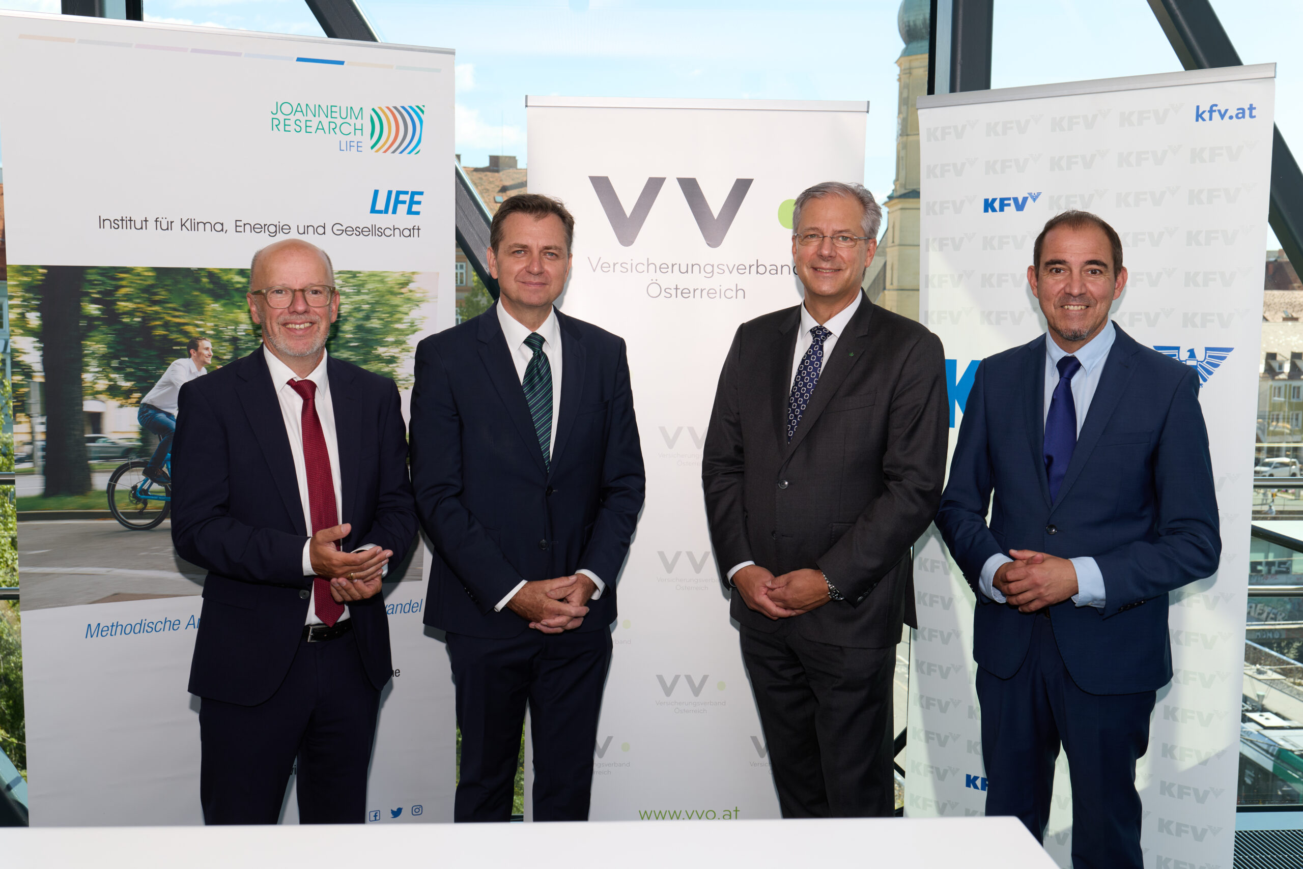 Vier Männer in Business-Kleidung stehen lächelnd vor Bannern von Joanneum Research LIFE, Versicherungsverband Österreich, KFV.at und der langen Tafel Graz in einem modernen Glasgebäude mit Stadtblick.