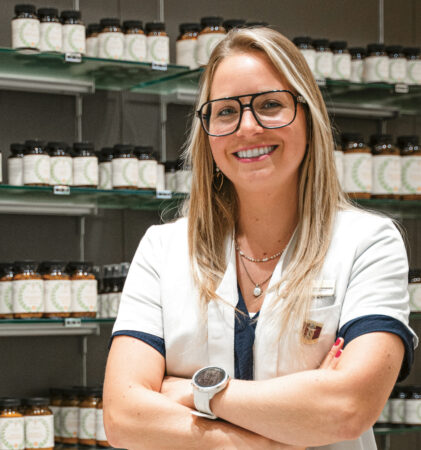 Eine lächelnde Frau in einem weißen Kittel mit Brille steht mit verschränkten Armen vor Regalen voller Gläser und Flaschen, wahrscheinlich in einer Apotheke oder einem Reformhaus.