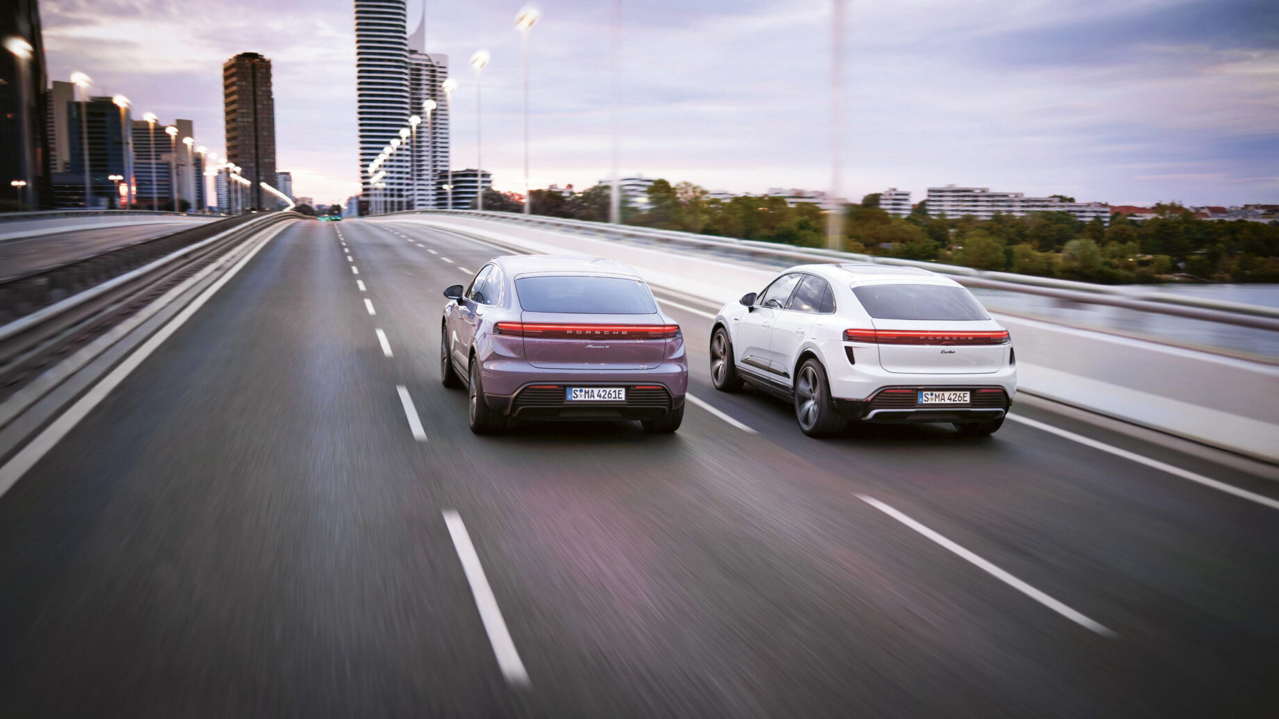 Zwei moderne Elektroautos fahren nebeneinander auf einer leeren mehrspurigen Brücke in der Abenddämmerung, mit städtischen Gebäuden und Bäumen im Hintergrund. Die Straße ist unscharf, was ihre Geschwindigkeit unterstreicht.