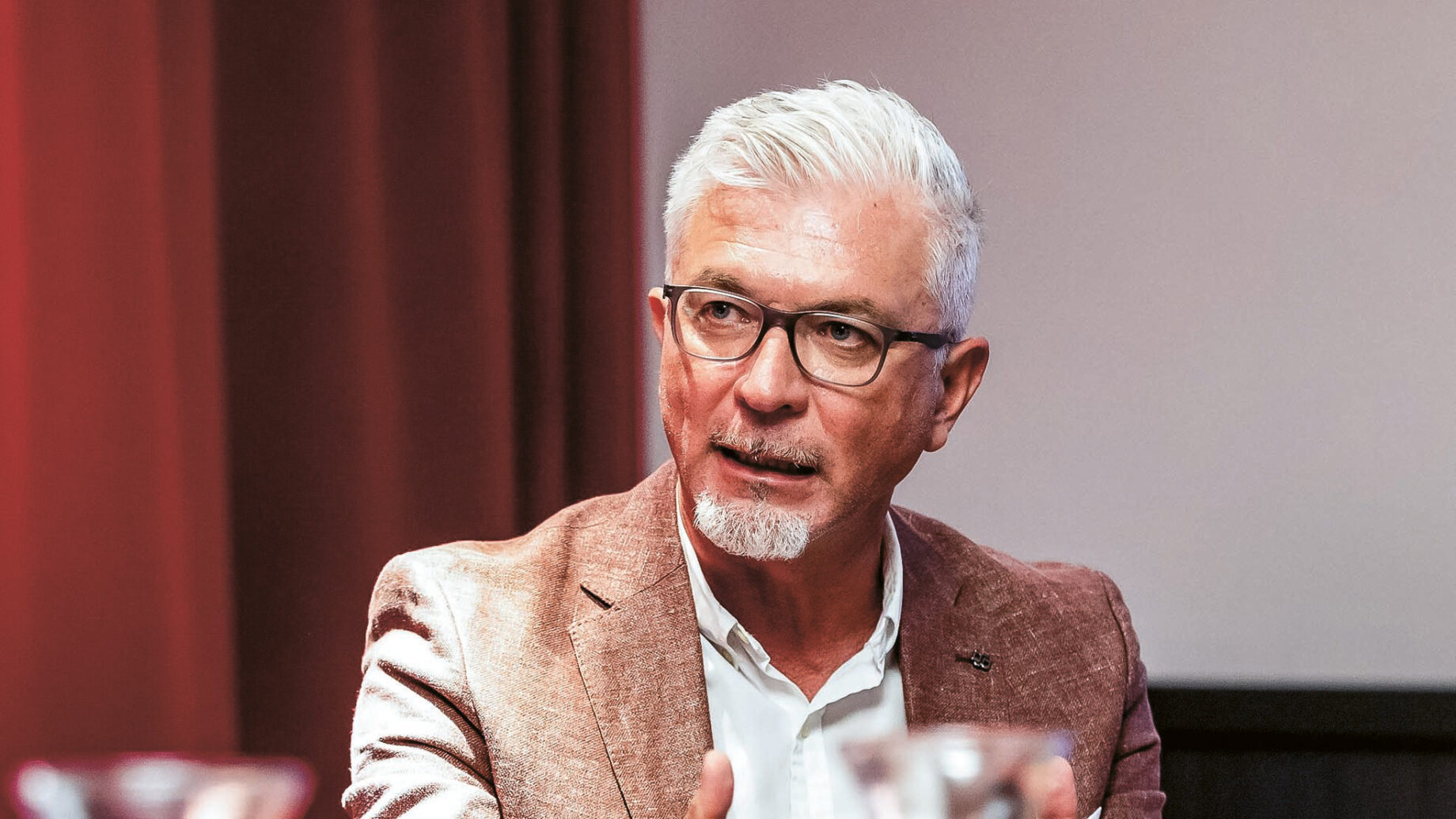 Ein Mann mit weißem Haar und Brille, brauner Jacke und weißem Hemd, sitzt an einem Tisch und gestikuliert mit den Händen. Vor ihm stehen Glaskrüge und Tassen, im Hintergrund ein roter Vorhang.