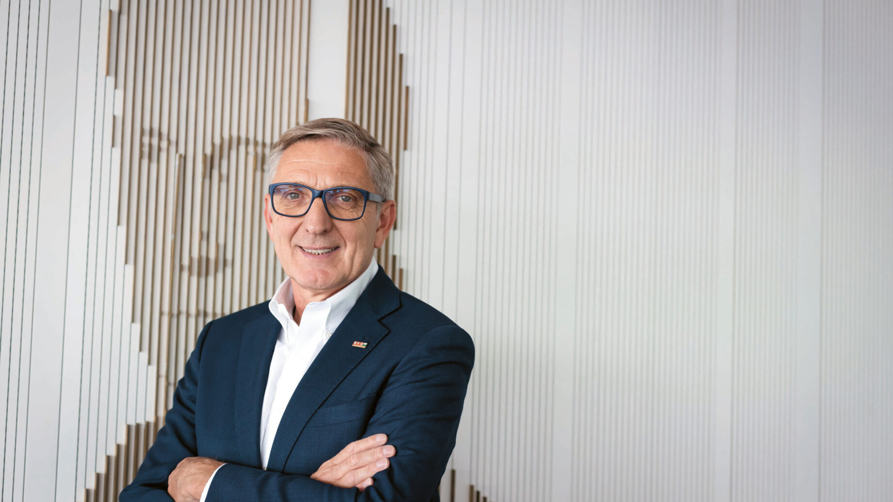 Ein Mann in einem dunklen Anzug und mit Brille steht mit verschränkten Armen lächelnd vor einer Wand mit einem strukturierten, abstrakten Porträtmotiv.