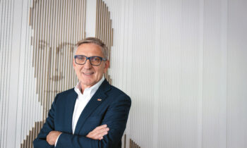 Ein Mann in einem dunklen Anzug und mit Brille steht mit verschränkten Armen lächelnd vor einer Wand mit einem strukturierten, abstrakten Porträtmotiv.