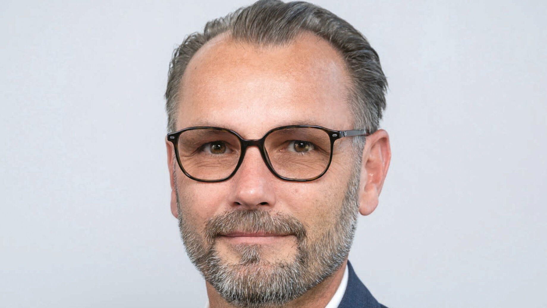 Ein Mann mit kurzen grauen Haaren und einem Bart, der eine schwarze Brille, ein weißes Hemd und eine dunkelblaue Anzugsjacke trägt, posiert vor einem schlichten hellgrauen Hintergrund.