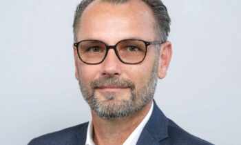 Ein Mann mit kurzen grauen Haaren und einem Bart, der eine schwarze Brille, ein weißes Hemd und eine dunkelblaue Anzugsjacke trägt, posiert vor einem schlichten hellgrauen Hintergrund.