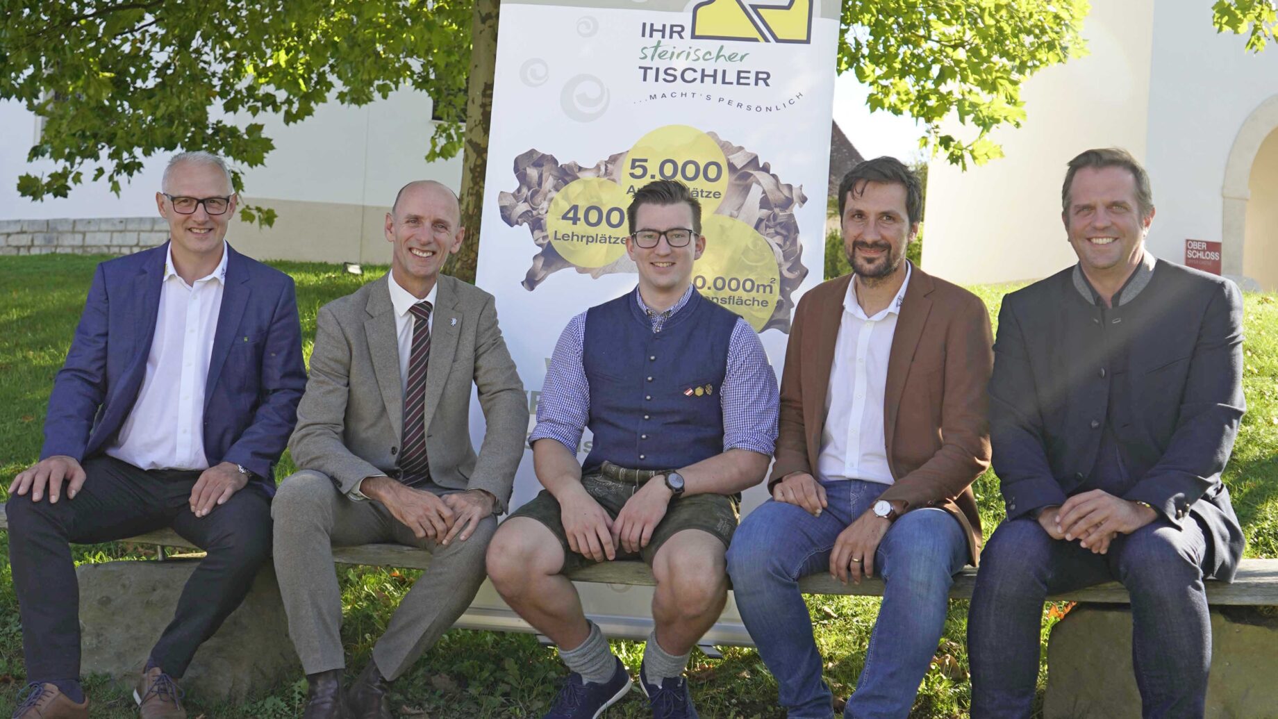 Fünf Männer in Geschäfts- und Freizeitkleidung sitzen auf einer Bank im Freien vor einem großen Baum und einem Werbebanner der Langen Tafel Graz und lächeln im hellen Sonnenlicht in die Kamera.