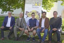 Fünf Männer in Geschäfts- und Freizeitkleidung sitzen auf einer Bank im Freien vor einem großen Baum und einem Werbebanner der Langen Tafel Graz und lächeln im hellen Sonnenlicht in die Kamera.