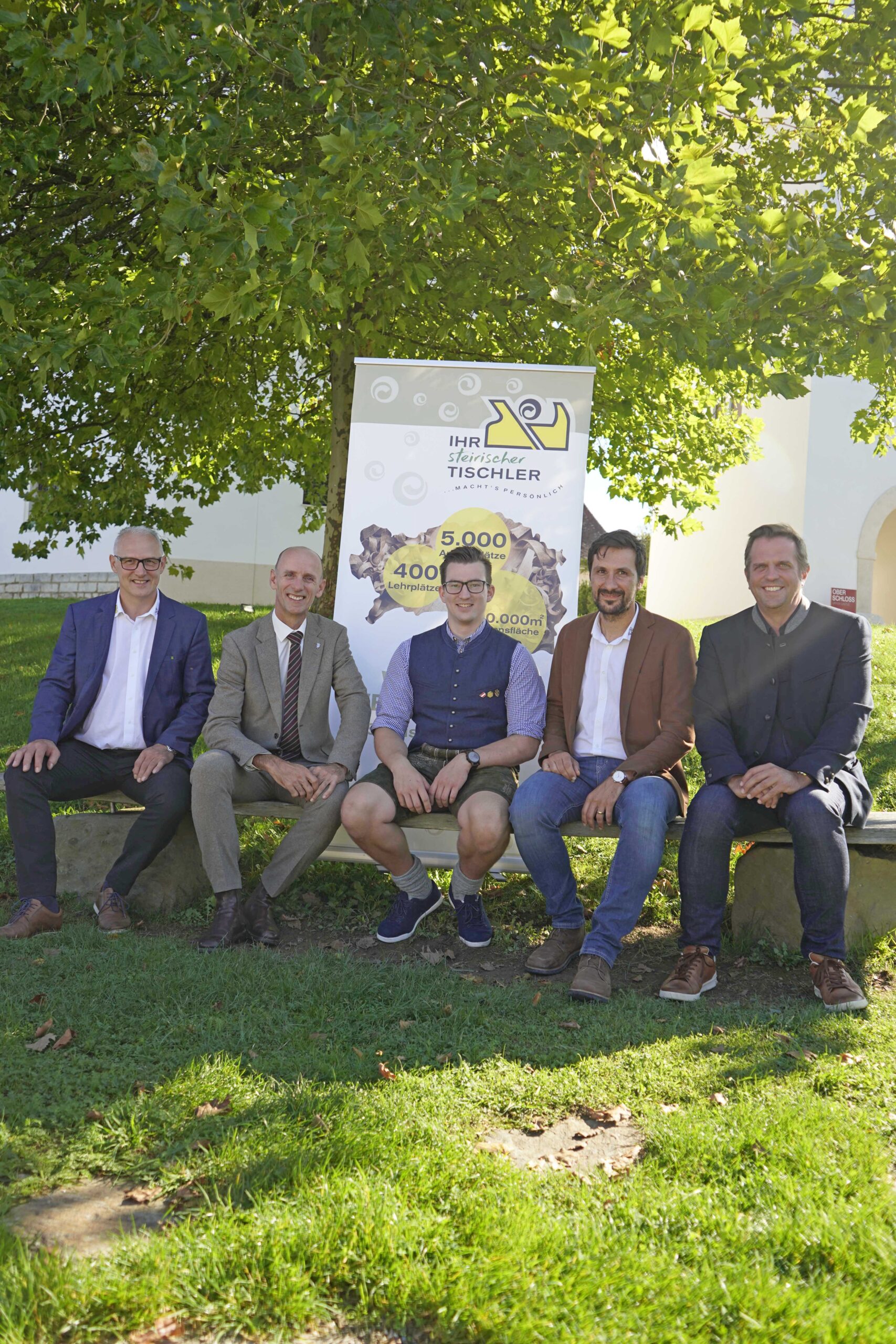 Fünf Männer in Geschäfts- und Freizeitkleidung sitzen auf einer Bank im Freien vor einem großen Baum und einem Werbebanner der Langen Tafel Graz und lächeln im hellen Sonnenlicht in die Kamera.