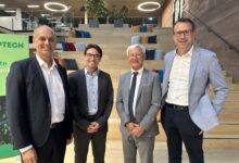 Vier Männer in Business-Kleidung stehen lächelnd vor einer Holztreppe mit bunten Kissen bei der langen Tafel Graz. Moderne, offene Büroräume sind zu sehen, mit kreisrunden Deckenleuchten und einem grünen Schild auf der linken Seite, auf dem "TECH.