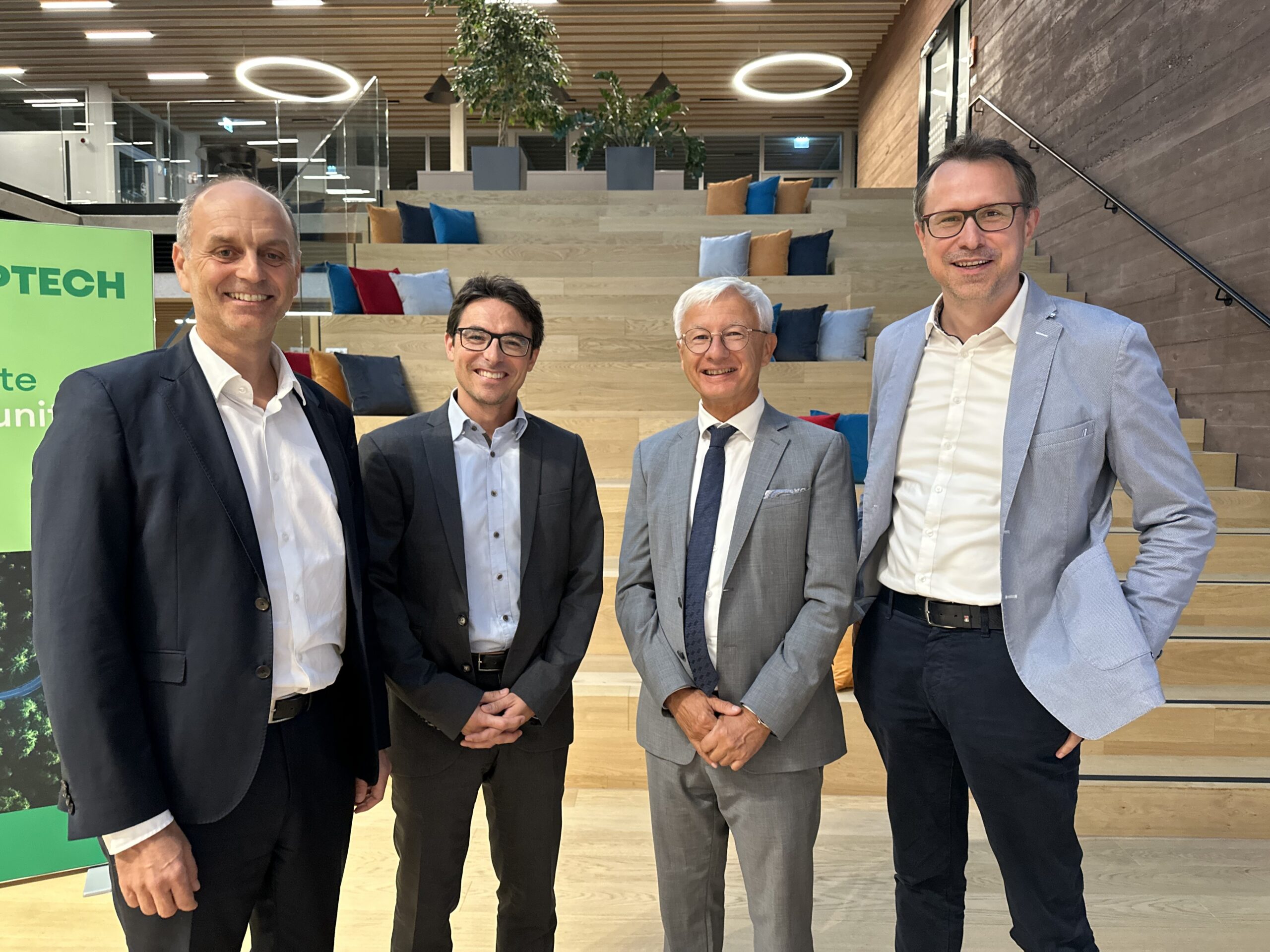 Vier Männer in Business-Kleidung stehen lächelnd vor einer Holztreppe mit bunten Kissen bei der langen Tafel Graz. Moderne, offene Büroräume sind zu sehen, mit kreisrunden Deckenleuchten und einem grünen Schild auf der linken Seite, auf dem "TECH.