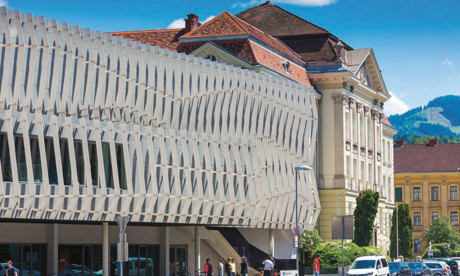 Ein modernes Gebäude mit einer einzigartigen, gewellten weißen Fassade steht neben einem älteren, klassizistischen Gebäude mit einem roten Ziegeldach. Auf dem Bürgersteig darunter sind Menschen und Fahrräder zu sehen, während im Hintergrund der blaue Himmel und die Berge zu sehen sind.