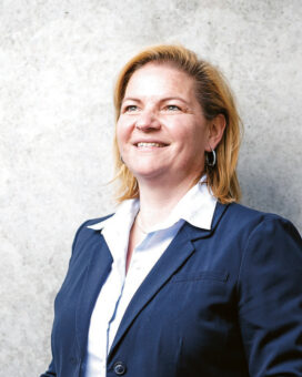 Eine Frau in einem marineblauen Blazer und weißem Hemd steht vor einer grauen Betonwand und lächelt selbstbewusst, die Hände in den Taschen.
