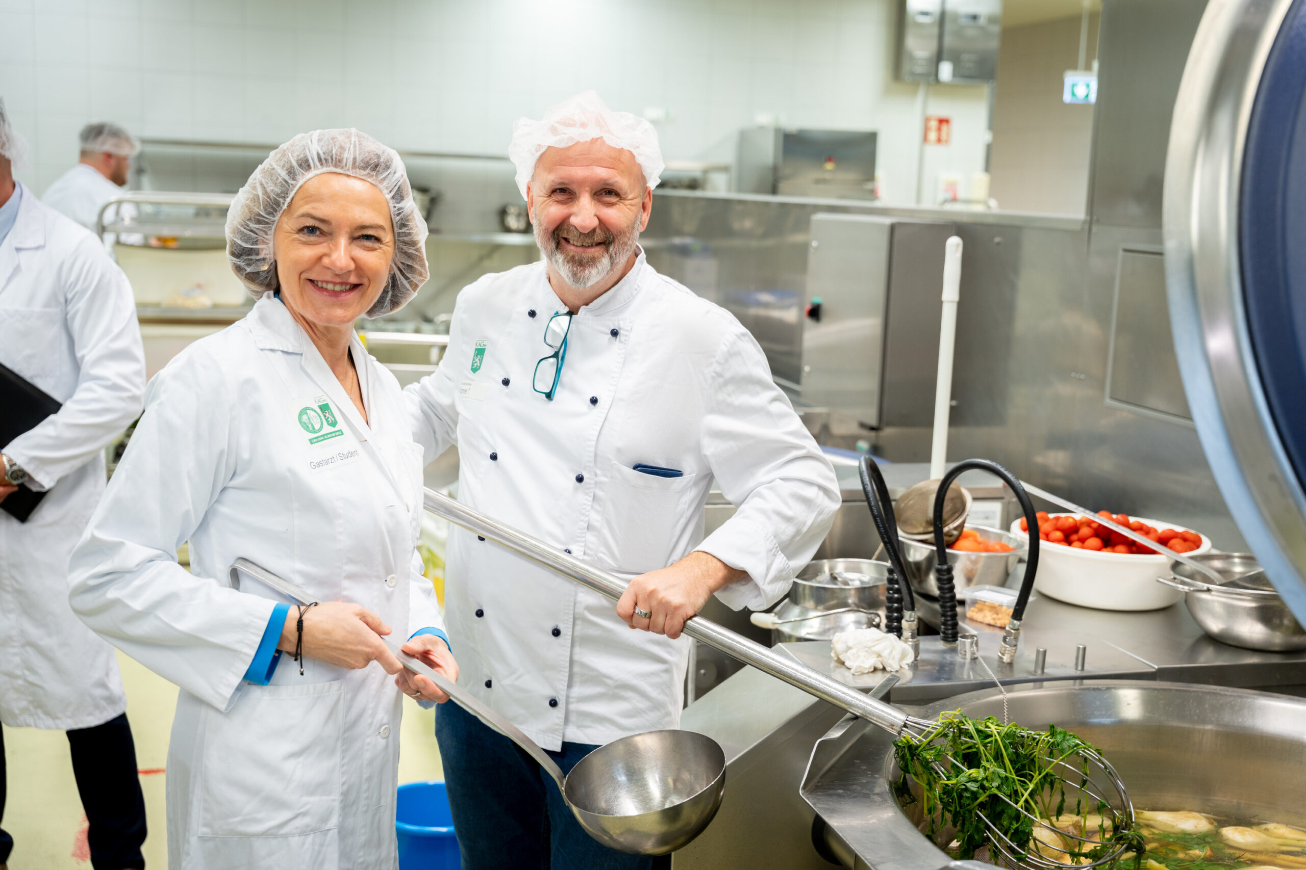 Zwei lächelnde Köche in weißen Uniformen und Haarnetzen stehen in einer Großküche und kochen mit frischen Kräutern in einem großen Topf. Edelstahlgeräte und Schüsseln mit Zutaten sind zu sehen - die Vorbereitungen für die lange Tafel Graz sind im Gange.