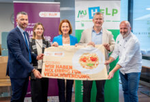 Fünf Personen stehen lächelnd in einem Raum und halten ein großes Schild mit einem Teller mit Lebensmitteln und einem deutschen Text über die Vermeidung von Lebensmittelabfällen bei der langen Tafel Graz. Banner im Hintergrund zeigen Logos und Botschaften von Organisationen.