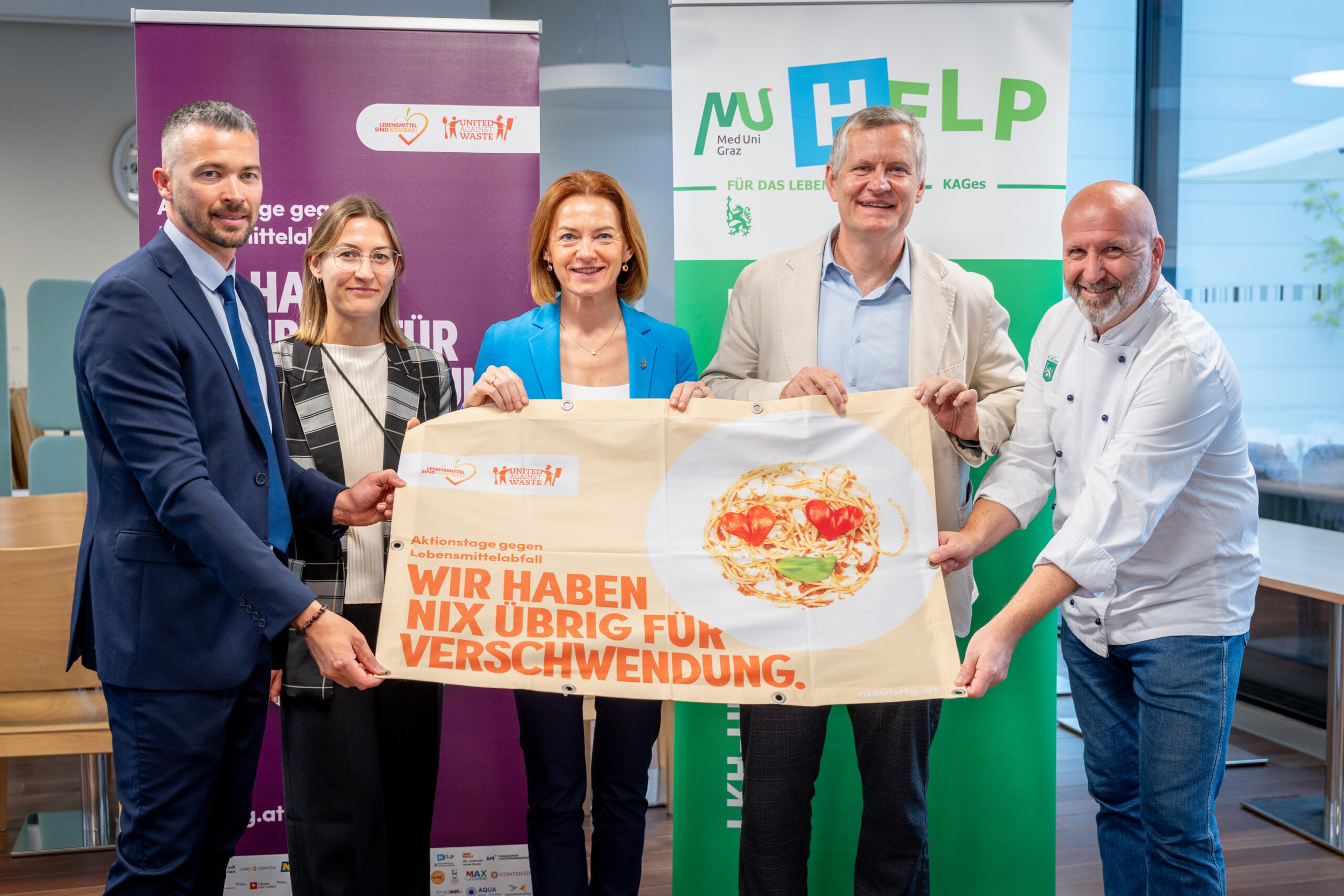 Fünf Personen stehen lächelnd in einem Raum und halten ein großes Schild mit einem Teller mit Lebensmitteln und einem deutschen Text über die Vermeidung von Lebensmittelabfällen bei der langen Tafel Graz. Banner im Hintergrund zeigen Logos und Botschaften von Organisationen.