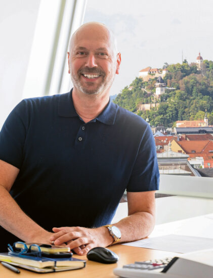 Ein lächelnder Mann mit Glatze und dunklem Polohemd sitzt an einem Schreibtisch mit Büchern, Papieren und einem Taschenrechner. Hinter ihm sind durch große Fenster eine Stadtlandschaft und ein Gebäude auf einem Hügel zu sehen.