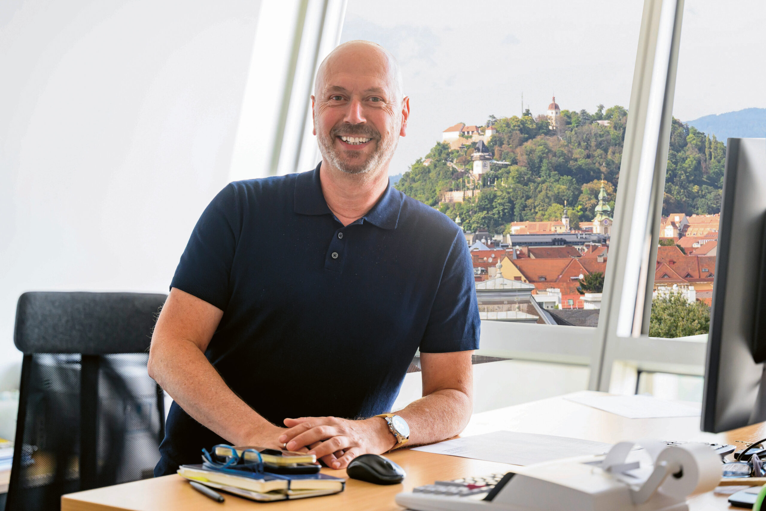 Ein lächelnder Mann mit Glatze und dunklem Polohemd sitzt an einem Schreibtisch mit Büchern, Papieren und einem Taschenrechner. Hinter ihm sind durch große Fenster eine Stadtlandschaft und ein Gebäude auf einem Hügel zu sehen.