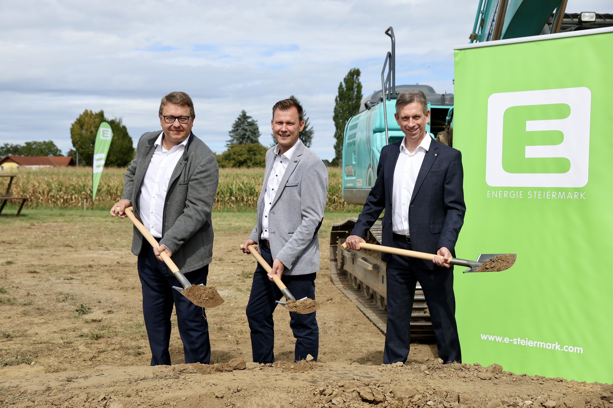 Drei Männer in Anzügen stehen im Freien und halten Schaufeln mit Erde in der Hand. Sie nehmen an einem Spatenstich teil, der neben einem grünen "Energie Steiermark"-Banner, Baumaschinen und Schildern für die lange Tafel Graz stattfindet.