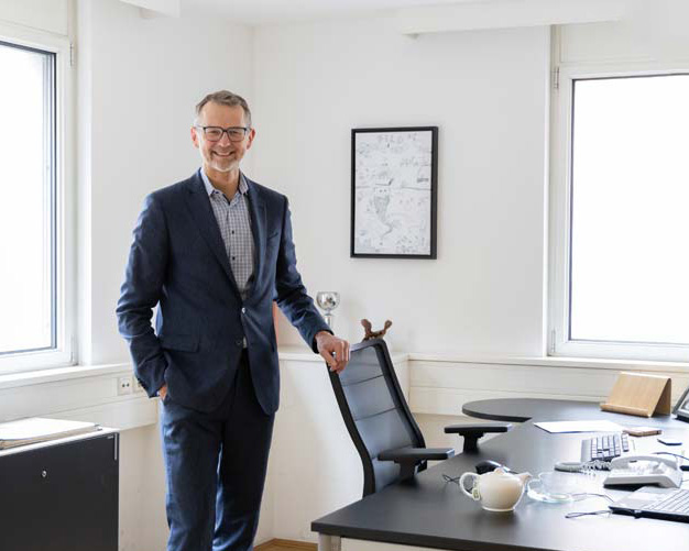 Ein Mann im Anzug steht lächelnd neben einem schwarzen Bürostuhl in einem hellen, modernen Büro mit großen Fenstern, einem Schreibtisch, einem Computer und einer Kaffeetasse.