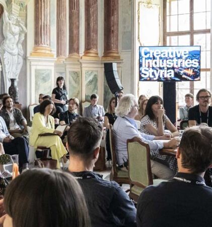 Eine große Gruppe von Menschen sitzt in einem schmucken Raum mit hohen Fenstern und Marmorwänden und hört aufmerksam einer Podiumsdiskussion zu. Zwei Bildschirme zeigen "Creative Industries Styria".