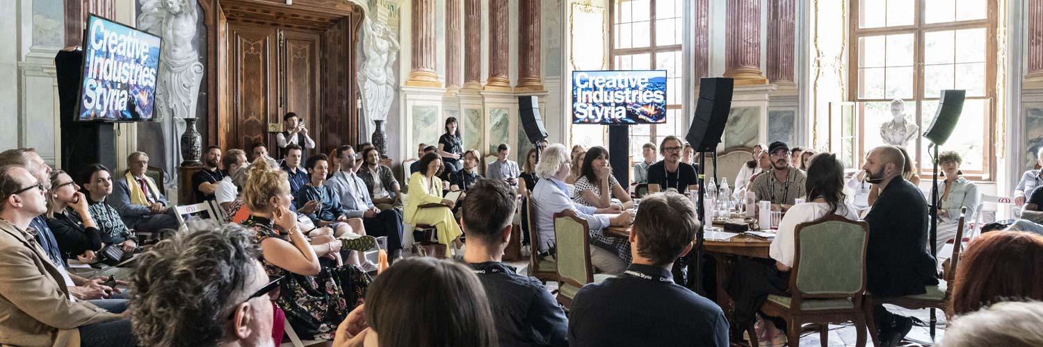 Eine große Gruppe von Menschen sitzt in einem schmucken Raum mit hohen Fenstern und Marmorwänden und hört aufmerksam einer Podiumsdiskussion zu. Zwei Bildschirme zeigen "Creative Industries Styria".